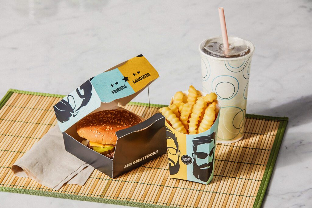 A view showcasing a burger in a fiber-based clamshell, French fry packaging, and cold cups in a quick service restaurant