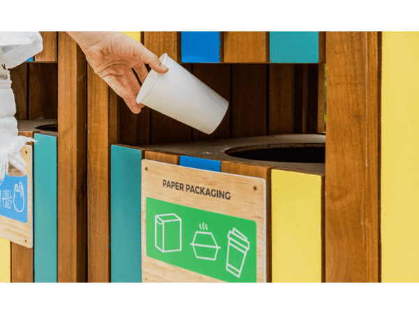 A person tosses a white paper cup into a colorful paper recycling bin outdoors.