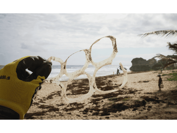A gloved hand holds a discarded six-pack plastic ring carrier collected from a sandy beach, with the ocean and shoreline in the background, illustrating the environmental impact of plastic packaging and the importance of plastic ring alternatives.